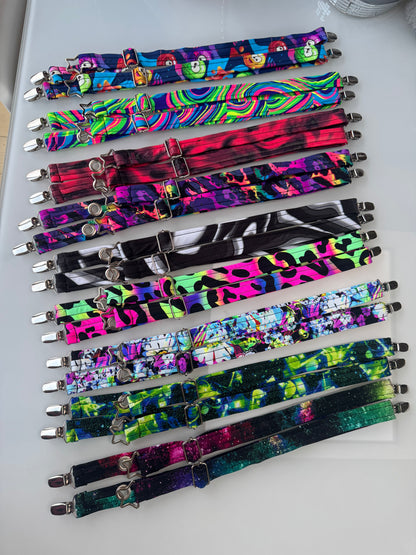 Nine pairs of colorful patterned Magic Utility Straps with silver metal clips are arranged vertically. The fabric patterns include various designs such as cartoon characters, wavy stripes, tie-dye, graffiti, and galaxy prints in vibrant neon and dark tones.