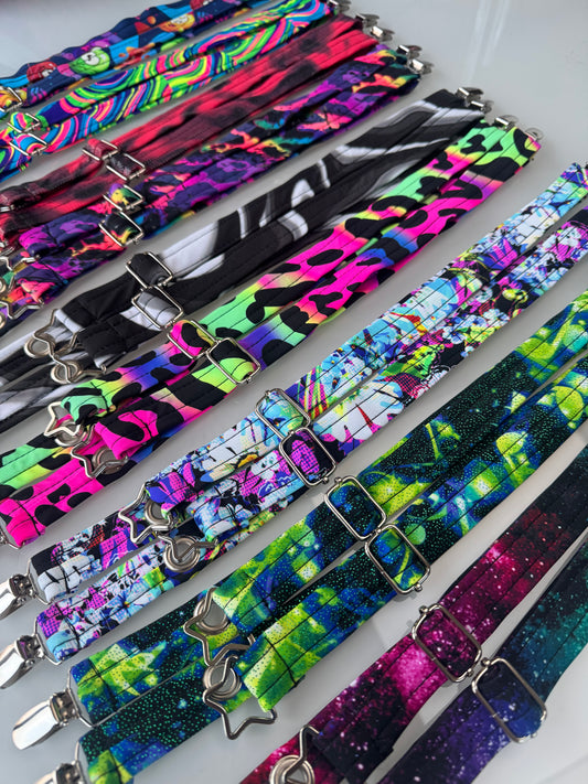 A collection of colorful jacket straps with silver metal buckles, arranged diagonally laid flat. The straps feature a variety of vibrant patterns including neon leopard, floral, abstract, and galaxy prints.